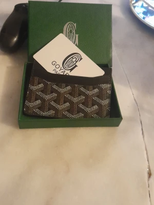  Svart/brun korthållare - Snygg svart/brun korthållare med det klassiska vita och svarta mönstret. Tillverkad i canvas med läderdetaljer och levereras i original grön ask. Perfekt för kort och sedlar, stilren och exklusiv design.