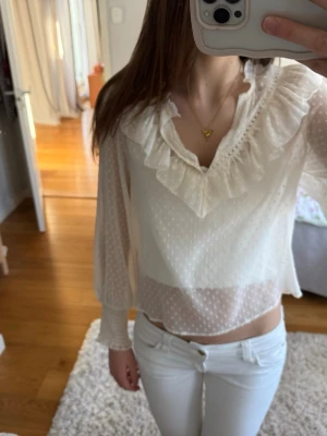 Vit blus med volang och prickigt mönster - Söt vit blus med transparent tyg och diskret prickigt mönster. Blusen har en djup v-ringning med volangdetaljer runt halsen och långa ärmar med smock vid ärmsluten. Perfekt för en trendig och romantisk look.
