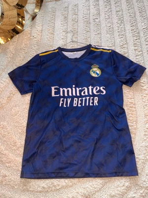Real Madrid blå matchtröja S - Snygg blå Real Madrid matchtröja från Adidas med gula detaljer på axlarna och klubbmärke på bröstet. Tillverkad i lätt och ventilerande polyester, perfekt för fotbollsträning eller sport. Tryck med 'Emirates Fly Better' på framsidan.