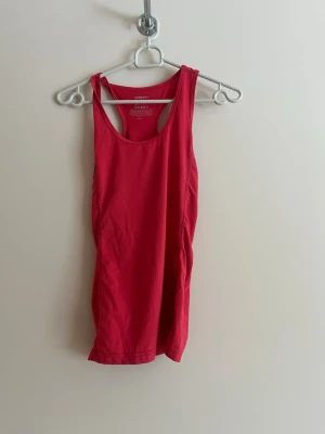 Röd linnetopp från Newbody - Snygg röd linnetopp från Newbody med klassisk racerback-design. Perfekt för träning eller sommarens varma dagar. Tillverkad i mjuk och stretchig polyester som sitter skönt mot kroppen. Enkel och stilren modell utan mönster eller extra detaljer.