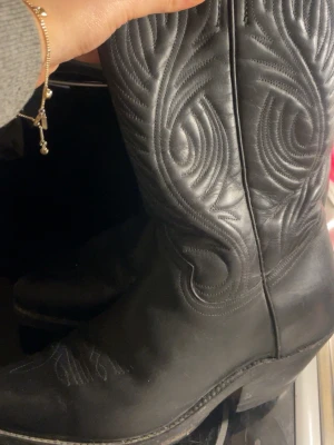Svarta cowboy boots i läder - Säljer ett par klassiska svarta cowboy boots i läder med snygga broderade detaljer längs skaftet. De har spetsig tå och stadig klack, perfekt för dig som vill ha en cool westernstil. Passar till jeans eller klänning för en trendig look.