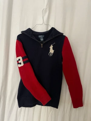 Mörkblå quarter zip tröja Polo Ralph Lauren - Stickad tröja i mörkblått med röda ärmar från Polo Ralph Lauren. Halvzip med hög krage, stor vit logga på bröstet och siffran 3 på ena ärmen. Ribbstickade muddar och sportig look. Perfekt för dig som gillar stil.