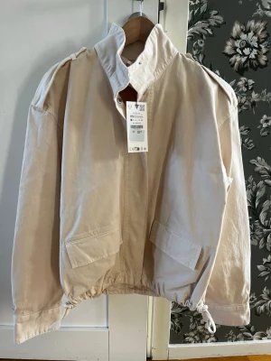 Zara cargojacka i storlek XS - Stilren beige vindjacka med hög krage och dragsko i nederkant. Jackan har två stora fickor framtill, axelklaffar och en avslappnad passform. Materialet är kraftigt bomullsliknande tyg som ger en cool och trendig look.