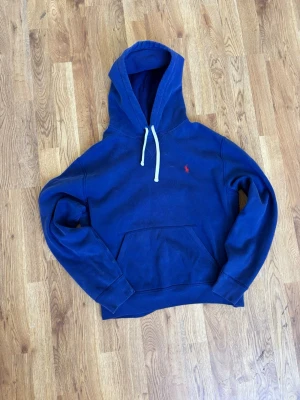 Blå hoodie från Polo Ralph Lauren - Mörkblå hoodie från Polo Ralph Lauren med klassisk huva och vita dragsnören. Tröjan har en känguruficka framtill och den ikoniska röda logotypen broderad på bröstet. Tillverkad i mjuk bomullsblandning med ribbade muddar vid ärmslut och nederkant.