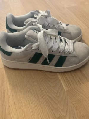 Adidas Campus sneakers i grå/vit och grönt - Säljer ett par Adidas Campus sneakers med grå-vit mocka och gröna detaljer. Skorna har klassiska vita snören, gröna ränder på sidorna och Adidas-logga på hälen. Köpta här på Plick, passade inte mig. Pris kan diskuteras!