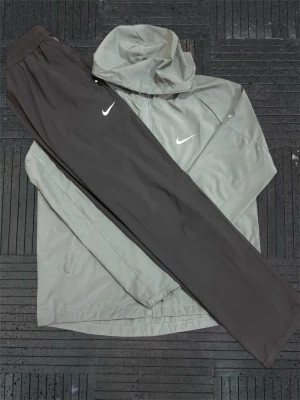 Nike träningsset jacka och byxor - Snyggt träningsset från Nike med en ljusgrå vindjacka med huva och dragkedja samt svarta träningsbyxor med resår i midjan. Båda plaggen har den klassiska vita Nike-loggan. Perfekt för löpning eller gym.