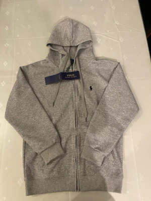 Grå hoodie från Polo Ralph Lauren - Klassisk grå hoodie från Polo Ralph Lauren med dragkedja framtill, justerbar huva med snören och ribbade muddar. Liten broderad logga på bröstet. Tillverkad i mjuk bomull för maximal komfort. Perfekt för en avslappnad och stilren look.