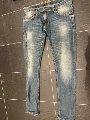 Blå slim fit jeans från Nudie Jeans - Säljer ett par blå slim fit jeans från Nudie Jeans med snygga slitningar och klassiska kontrastsömmar. Jeansen har fem fickor, gylf med dragkedja och Nudie-logga på bakfickan. Tillverkade i mjuk denim med stretch för skön passform. Passar 32!