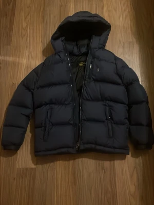 Svart pufferjacka från Polo Ralph Lauren - Säljer en svart pufferjacka från Polo Ralph Lauren med huva och dragkedja. Jackan har två stora fickor framtill och en polo logga på bröstet. Perfekt för kalla dagar och riktigt skön att bära.