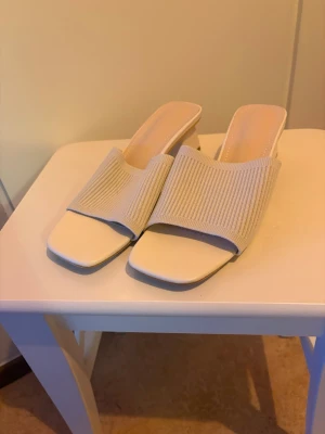 Beige ribbade slip-in klackar - Säljer ett par helt oanvända Snygga slip-in klackar i beige med ribbad textil över foten och fyrkantig tå. Skon har en låg, bred klack och öppen häl för en modern vibe. Perfekt till sommarens outfits och lätta att matcha med olika stilar.