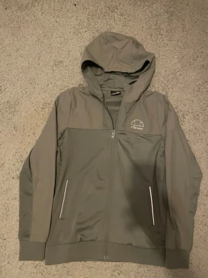 Grå vindjacka från Ellesse - Grå vindjacka från Ellesse med huva och hel dragkedja framtill. Två fickor med dragkedja och diskret Ellesse-logga på bröstet. Tillverkad i lätt och vindtätt material, perfekt för blåsiga dagar. Ribbade muddar vid ärmslut och nederkant för extra komfort. Pris ej hugget i sten . Skriv för frågor!