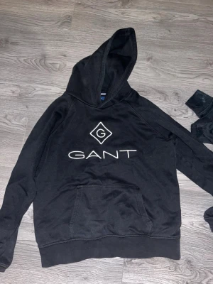 Svart hoodie från GANT med logga - Snygg svart hoodie från GANT med stor vit logga och emblem på bröstet. Klassisk känguruficka framtill och huva. Perfekt för en avslappnad och trendig stil. Tillverkad i mjukt material för extra komfort. Säljer pga att den är för liten för mig. Storlek 176cm 16 years old. Hör av dig vid frågor