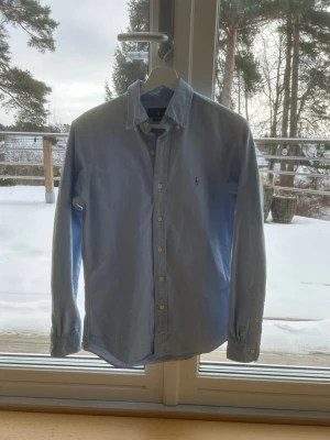Ljusblå skjorta från Ralph Lauren - Klassisk ljusblå skjorta från Ralph Lauren med button-down krage och broderad logga på bröstet. Skjortan har långa ärmar, vita knappar och är tillverkad i mjuk bomull. Perfekt för en clean och stilren look. Skjortan är i ny skick!! Ny pris 2200kr mitt pris 399kr