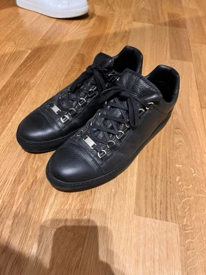 Balenciaga Arena Low - Stilrena svarta sneakers från Balenciaga i mjukt läder med svarta skosnören och metallöglor. Klassisk låg modell med rund tå och diskret logga på plösen. Perfekta för dig som gillar minimalistisk och lyxig design.