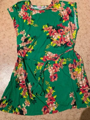 Grön blommig klänning från Indiska - Grön kortklänning från Indiska med färgglatt blommönster i rosa, rött och gult. Klänningen har korta ärmar, rund halsringning och ett knytband i midjan för en snygg passform. Perfekt för varma dagar och somriga tillfällen. Lite hål bak