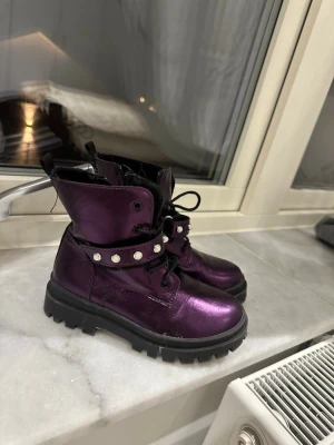 Lila metallic kängor med pärldetaljer - Snygga kängor i metallic-lila med grov sula och svarta snören. Dekorerade med ett band med pärlor runt ankeln för extra attityd. För barn.