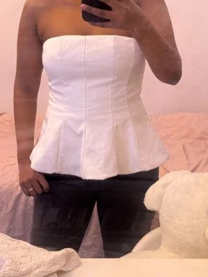 Vit korsett-topp med peplum - Säljer en snygg vit korsett-topp med peplumformad nederkant. Toppen är axelbandslös och har markerade sömmar som ger en strukturerad look. Perfekt för dig som vill ha en stilren och trendig topp till din outfit.