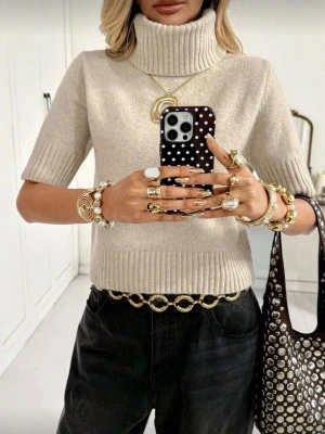 Beige stickad polotröja kort ärm - Trendig beige stickad polotröja med korta ärmar och bred ribbad kant vid ärmslut och nederkant. Hög, vikt polokrage ger en cool look. Perfekt att styla med jeans eller kjol för en avslappnad men snygg outfit.