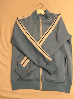 Ljusblå zip hoodie från Palm Angels - Snygg ljusblå zip hoodie från Palm Angels med vita och svarta ränder längs ärmarna. Dragkedja framtill, ribbade muddar och fickor på sidorna. Tillverkad i ett mjukt, sportigt material och har Palm Angels-logga tryckt på bröstet.