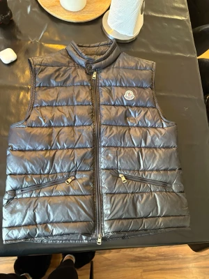 Svart dunväst från Moncler - Snygg svart dunväst från Moncler med quiltad design och hög krage. Västen har två dragkedjeförsedda fickor framtill och en Moncler-logga på bröstet. Perfekt för lager-på-lager och ger en clean look till din outfit.