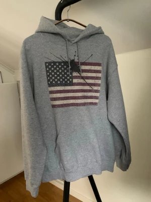 Grå hoodie med USA-flagga från Concrete Ink. - Säljer en grå hoodie med tryck av en amerikansk flagga på bröstet. Hoodien har huva med dragsko och en avslappnad passform. Perfekt för dig som gillar streetwear och vill sticka ut lite extra. Hör av dig vid minsta fundering eller fråga och pris kan diskuteras! 🤩🙌