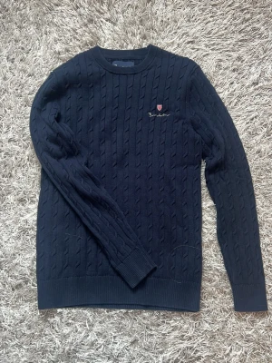 Mörkblå kabelstickad tröja Barbour - Snygg mörkblå kabelstickad tröja  med rund halsringning och diskret broderad logga på bröstet. Tröjan har långa ärmar och ribbade muddar vid ärmslut och nederkant. 