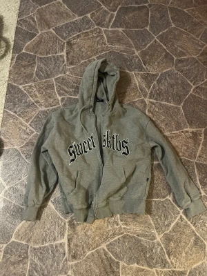 Grå hoodie med tryck Sweet Sk8tbs - Grå ziphoodie med svart tryck 'Sweet Sk8tbs' i gotisk stil på bröstet. Klassisk modell med huva och fickor framtill. Tillverkad i mjukt sweatshirtmaterial, perfekt för en avslappnad streetwear-look.