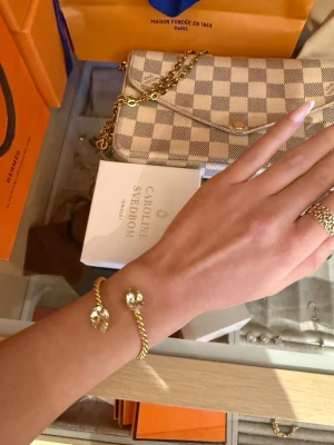 Guldarmband Caroline Svedbom - Snyggt armband i guldton från Caroline Svedbom med tvinnad design och två stora, gnistrande pärlor i ändarna. Armbandet är öppet och justerbart, perfekt för att ge en lyxig touch till din outfit.