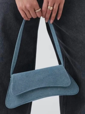 Blå denim handväska Gina Tricot - Trendig handväska i ljusblå denim från Gina Tricot. Väskan har en avlång, mjukt trekantig form med lock och magnetknäppning. Ett axelband gör den enkel att bära och insidan har en liten ficka. Perfekt accessoar för en cool och avslappnad look.