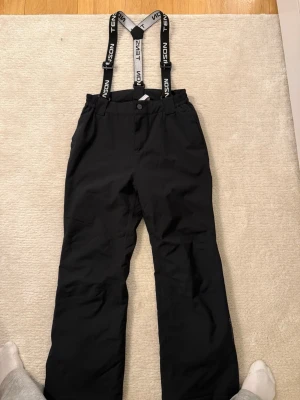 Svarta täckbyxor med hängslen Tenson - Säljer ett par svarta täckbyxor från Tenson med justerbara hängslen och raka ben. Byxorna har knapp- och dragkedjestängning, samt praktiska fickor framtill. Perfekta för vinterns alla äventyr i snön.