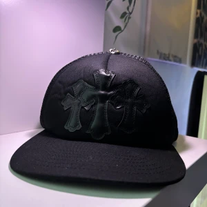 Svart keps från Chrome Hearts - Säljer en svart keps från Chrome Hearts med tre svarta kors i läder framtill och broderad logga bak. Kepsen har böjd skärm och justerbar snapback. Materialet är en mix av tyg och mesh för skön ventilation.