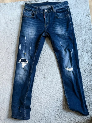 Blå slitna jeans från Dsquared2 - Säljer ett par blå jeans från Dsquared2 med coola slitningar och hål på benen. Jeansen har klassisk femficksmodell, knappgylf och tydliga slitdetaljer för en trendig look. Perfekta för dig som gillar streetstyle och vill sticka ut.