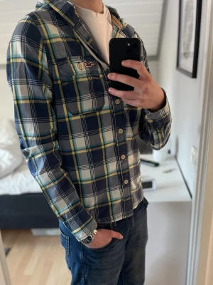 Rutig blå skjorta med huva Hollister - Säljer nu en snygg rutig skjorta från Hollister i blå, vit och gul med huva och snörning. Skick 10/10, dvs ser helt ny ut. Modellen på bilden är 183 och väger 67 kilo. Skriv om ni har några mer funderingar😊