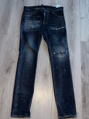 Mörkblå jeans av märket Dsquared2 - Hej! Säljer nu mina mörkblå jeans från Dsquared2 med slitningar, färgstänk och trasiga detaljer på benen. Smal passform och klassisk femficksmodell. Säljer de eftersom jag inte använder de så mycket, skicket är mycket bra. Vid intresse är det bara att höra av sig. Storleken är 48, vilket motsvarar ungefär M