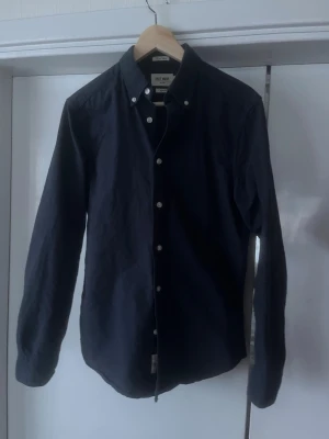 Mörkblå skjorta från East West - Snygg mörkblå skjorta från East West med klassisk button-down krage och vita knappar. Skjortan har lång ärm och en normal passform, perfekt att bära öppen över t-shirt eller stängd. Tillverkad i mjuk bomull för en bekväm känsla.