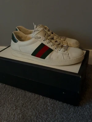 Gucci vita sneakers med röd/grön rand - Säljer ett par klassiska vita sneakers från Gucci i läder med den ikoniska röd-gröna randen på sidan och mörkgrön detalj vid hälen. Skorna har lågt skaft och vita snören. Perfekta för dig som vill ha en stilren och lyxig look.