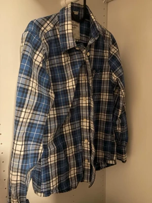 Blårutig skjorta Abercrombie & Fitch - Klassisk blårutig skjorta från Abercrombie & Fitch med lång ärm och knappar framtill. Skjortan har en avslappnad passform och är tillverkad i mjuk bomull. Perfekt för en casual look med tidlös stil. Använd 2-3 gånger nästan ny