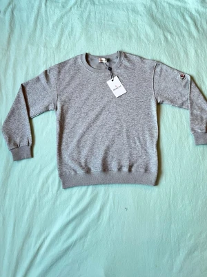 Grå sweatshirt från Moncler - Snygg grå sweatshirt från Moncler med rund halsringning och ribbade muddar vid ärmslut och nederkant. Tröjan har en diskret Moncler-logga på ena ärmen och är tillverkad i mjuk bomullsblandning. Perfekt för en clean och stilren look.