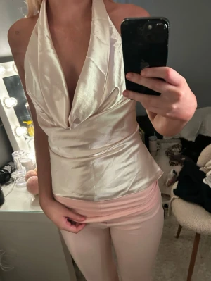 Champagnefärgad öppen rygg i satin - Snygg champagnefärgad halterneck topp i glansigt satinmaterial. Toppen har djup urringning och omlott-drapering framtill som ger en elegant look.  