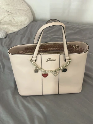 Beige handväska från Guess med kedja - Snygg beige handväska från Guess i konstläder med två handtag och guldfärgad kedjedetalj med berlocker framtill. Väskan har dragkedja upptill och ett mönstrat innerfoder med flera fack för smart förvaring. Perfekt för dig som gillar stilrena accessoarer.