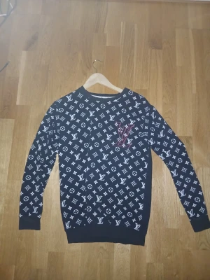 Louis Vuitton  - Svart sweatshirt från Louis Vuitton med vitt monogramtryck över hela framsidan och ärmarna. Tröjan har rund halsringning, ribbade muddar och en stor LV-logga i rött på bröstet. Perfekt för dig som gillar streetwear och exklusiva märken.