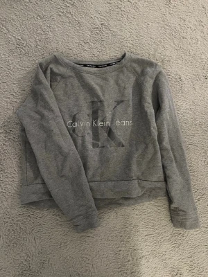 Grå sweatshirt från Calvin Klein Jeans - Säljer en stilren grå sweatshirt från Calvin Klein Jeans med stort CK-tryck och logga på bröstet. Tröjan har rund halsringning och långa ärmar. Tillverkad i mjukt bomullsmaterial som är skönt mot huden. Perfekt för en avslappnad och trendig look. Den är lite croppad och märket är också lite holografiskt. 