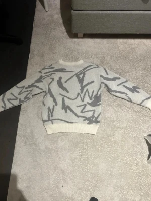 stickad tröja - Snygg stickad tröja i vitt med abstrakt grått mönster. Tröjan har rund halsringning, långa ärmar och ribbade muddar vid ärmslut och nederkant. Perfekt för dig som gillar en modern och unik stil.