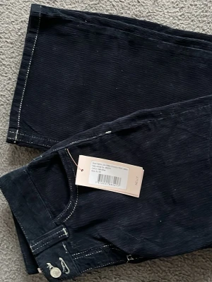 Mörkblå manchesterbyxor med detaljer - Säljer ett par mörkblå från Nelly med boot cut, low waist och kontrastsömmar. Baksidan har fickor med vita dekorativa sömmar i zigzag-mönster. Byxorna har knapp och dragkedja framtill samt hällor för skärp. Jeansen är helt ny och oanvända då de är för små. Är öppen för pris förslag. de är i storlek 34💕