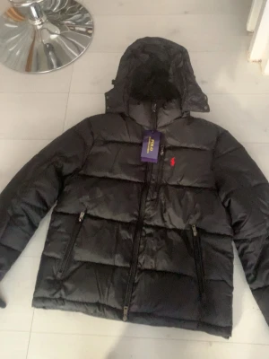 Svart dunjacka från Polo Ralph Lauren - Snygg svart dunjacka från Polo Ralph Lauren med huva och dragkedja framtill. Jackan har två framfickor med dragkedja och den klassiska röda loggan broderad på bröstet. Perfekt för kalla vinterdagar och riktigt stilren look.