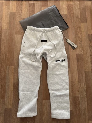 Grå Essentials Fear of God mjukisbyxor - Snygga ljusgrå mjukisbyxor broderad logga på benet. Byxorna har resår i midjan, långa dragsnören och sidofickor. Tillverkade i mjuk bomullsblandning för maximal komfort. Perfekta för en chill och trendig look.