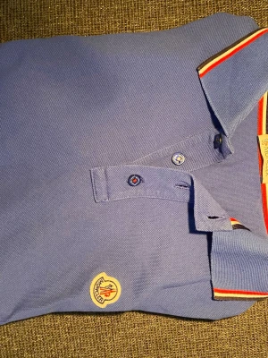 Blå Moncler pikétröja med röd detalj - Säljer en blå pikétröja från Moncler med klassisk krage och knappar framtill. Tröjan har röda, vita och mörkablåa  detaljer på kragen samt Moncler-logga på bröstet. Tillverkad i bomull för en skön och luftig känsla. Köpt i USA, kvitto finns