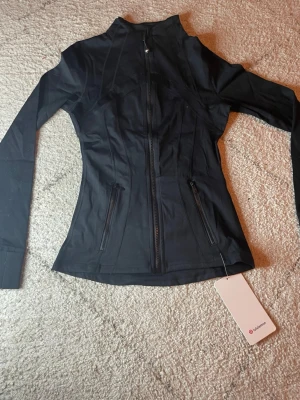  Lululemon träningsjacka - Slimmad svart träningsjacka från Lululemon med hel dragkedja framtill och två dragkedjeförsedda fickor. Tillverkad i stretchigt funktionsmaterial som andas, perfekt för löpning eller gym. Hög krage och långa ärmar för extra komfort.