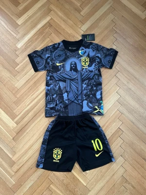 Brasilien Neymar Jr. matchställ Nike - Snyggt svart matchställ från Nike med Brasilien-tema och Neymar Jr. nummer 10. Tröjan har ett detaljerat tryck med Kristusstatyn och brasilianska symboler, samt gula loggor och tryck. Shortsen är svarta med gula detaljer och Brasilien-emblem.