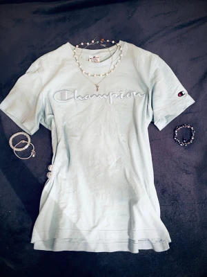 Ljusblå Champion t-shirt med snörning - Snygg ljusblå t-shirt från Champion med broderad logga framtill. T-shirten har korta ärmar och en relaxed passform. Perfekt för dig som gillar streetwear och vill sticka ut lite extra. Storlek xs men passar större i storleken.